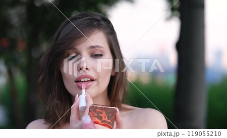 Close-up portrait of a beautiful woman who paints her lips with lipstick outdoors, looking into a pocket mirror. She makes a sexy face. 119055210