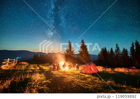 Group of friends hikers resting around campfire, surrounded by illuminated tents under stunning starry sky. Milky Way stretches above, creating magical atmosphere in peaceful mountain wilderness. Group of friends hikers resting around campfire, surrounded by illuminated tents under stunning starry sky. Milky Way stretches above, creating magical atmosphere in peaceful mountain wilderness. 119059892
