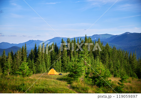 Bright yellow tourist tent on grassy hillside, surrounded by lush green pine trees. Breathtaking view of distant blue mountains under clear sky, creating idyllic and serene camping spot. Bright yellow tourist tent on grassy hillside, surrounded by lush green pine trees. Breathtaking view of distant blue mountains under clear sky, creating idyllic and serene camping spot. 119059897