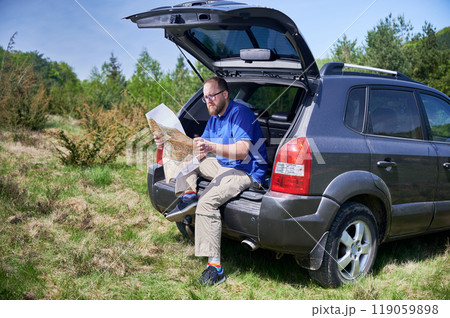 Man sits on open trunk of parked SUV in grassy, forested area, intently studying large map. Tourist navigating or planning route. Man sits on open trunk of parked SUV in grassy, forested area, intently studying large map. Tourist navigating or planning route. 119059898