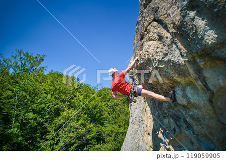 Aerial view of male rock climber ascending rugged limestone cliff with harness and rope for safety. Sportsman climbing on vertical large boulder at Dobvush Rocks in Carpathian mountains, Ukraine. Aerial view of male rock climber ascending rugged limestone cliff with harness and rope for safety. Sportsman climbing on vertical large boulder at Dobvush Rocks in Carpathian mountains, Ukraine. 119059905