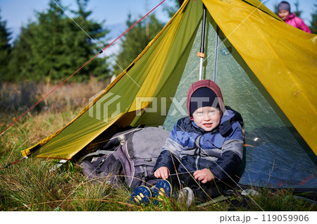 Happy young child sits at entrance of bright yellow tent in outdoor setting, wearing warm jacket and hat. Background features greenery and trees, suggesting camping trip in forested area. Happy young child sits at entrance of bright yellow tent in outdoor setting, wearing warm jacket and hat. Background features greenery and trees, suggesting camping trip in forested area. 119059906