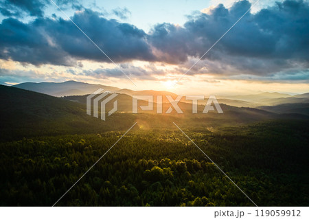 Aerial view breathtaking sunset over forest. Sun's rays break through dramatic clouds, casting golden glow across rolling hills and valleys. Distant mountains creating serene and picturesque scene. 119059912