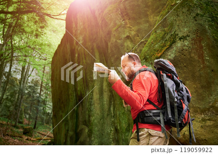 Bearded tourist man looks puzzled, studies map while stands in front of large rock in dense forest. Traveler with grey backpack and glasses, trying to figure out his direction. 119059922