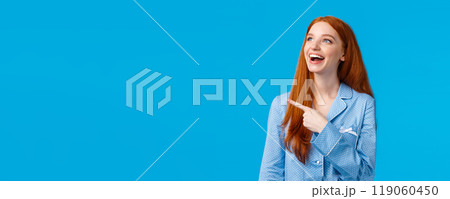 Delighted and entertained good-looking caucasian redhead girl in nightwear, wearing pyjama to sleep, pointing looking upper left corner amused, laughing happily, standing blue background 119060450