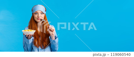Morning, lifestyle and women concept. Pretty cute redhead, foxy girl with long red hair waking-up early, prepare breakfast, biting spoon and smiling eating cereals, standing blue background 119060540
