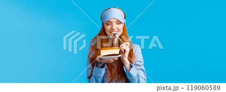 Girl feeling satisfaction eating sweet delicious dessert in morning. Attractive cute woman with red hair in nightwear and sleep mask, close eyes biting tasty cake, holding spoon close eyes 119060589