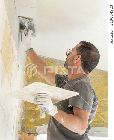Craftsman in dirty gloves and gray shirt spreading plaster with a metal float over a ceiling Craftsman in dirty gloves and gray shirt spreading plaster with a metal float over a ceiling 119064523