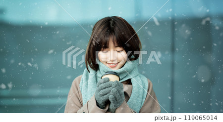 雪の降る冬の街角で暖かい飲み物を飲む女性 119065014