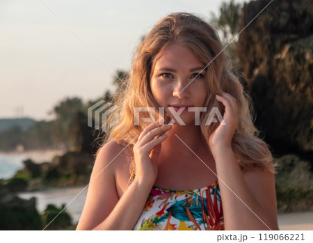 Portrait of a young woman with wavy blonde hair stands at the beach at the sunset. 119066221