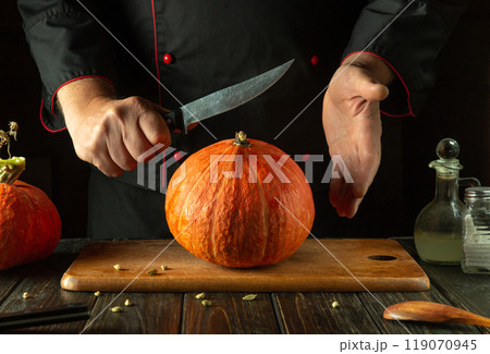 Preparing to cut a large orange pumpkin for a fruit menu. American cuisine or fruit diet concept. Knife in the chef hand Preparing to cut a large orange pumpkin for a fruit menu. American cuisine or fruit diet concept. Knife in the chef hand 119070945
