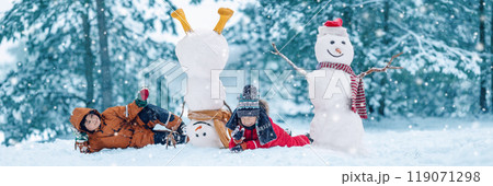 Two boys in warm clothing lying on the snow near snowmen in natural park. 119071298