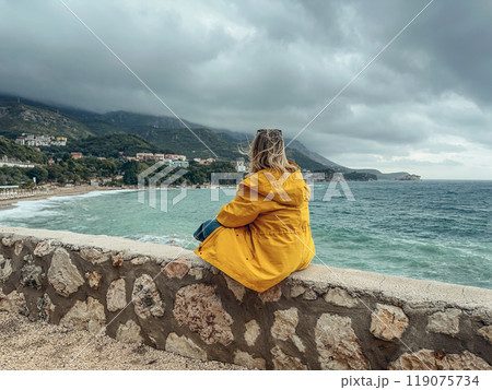 A woman in a yellow windbreaker looks out from a cliff at the sea A woman in a yellow windbreaker looks out from a cliff at the sea 119075734