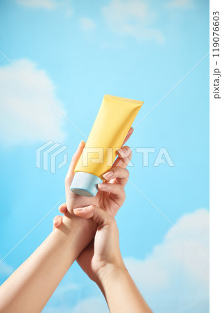 Under a blue sky with a few clouds and strong light, a hand is holding the wrist of another hand holding an unbranded yellow tube of cream with a blue cap, ideal for illustrating skincare content. Under a blue sky with a few clouds and strong light, a hand is holding the wrist of another hand holding an unbranded yellow tube of cream with a blue cap, ideal for illustrating skincare content. 119076603