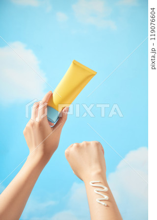 An unbranded yellow tube with a blue cream cap is held by a model, who is wearing a smear of cream on wrist, suitable for promoting a skincare product. There are clouds and a blue sky in the backdrop. 119076604