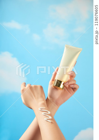 The model hands rub together, squeezing a zigzag cream streak and holding a beige tube cream mockup with a copper gold cap in a blue summer scene for sunscreen advertising photo templates. 119076605