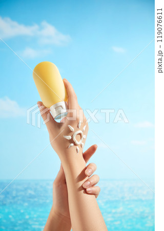 A female model hands are gently clasped together and holding a tube of unlabeled lemon yellow cream. On the back of her hand is a sun-shaped cream mark, suitable for advertising images for sunscreen. 119076611