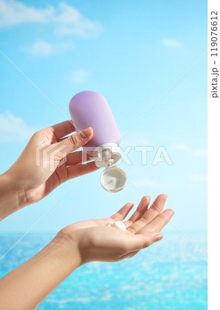 The backdrop of a sunny blue sky and sea features a female model hands squeezing an unbranded purple bottle of cream into a hand, which is perfect for a sunscreen advertisement image. The backdrop of a sunny blue sky and sea features a female model hands squeezing an unbranded purple bottle of cream into a hand, which is perfect for a sunscreen advertisement image. 119076612