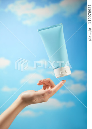 A female model hand is gently preparing to touch an unbranded blue cream tube with a white cap floating in the bright blue sky, a sample image for skincare advertisements. 119076621