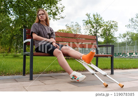 Casual-dressed female adolescent with cast on leg appears disheartened while seated on bench in summer park, conveying emotional and physical challenges during recovery. medical, disability, lifestyle Casual-dressed female adolescent with cast on leg appears disheartened while seated on bench in summer park, conveying emotional and physical challenges during recovery. medical, disability, lifestyle 119078385