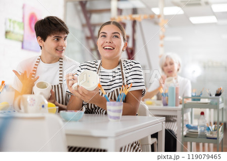 Young couple joyfully talking and painting ceramic mugs in pottery workshop 119079465