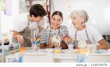 Senior woman assisting happy guy and girl painting ceramic mugs in workshop Senior woman assisting happy guy and girl painting ceramic mugs in workshop 119079479
