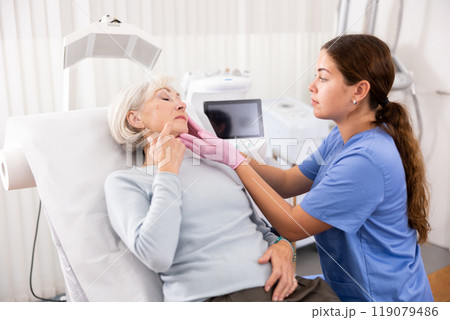 Mature woman lying on couch during consultation with female beautician, checking her skin before medical intervention in esthetic clinic Mature woman lying on couch during consultation with female beautician, checking her skin before medical intervention in esthetic clinic 119079486
