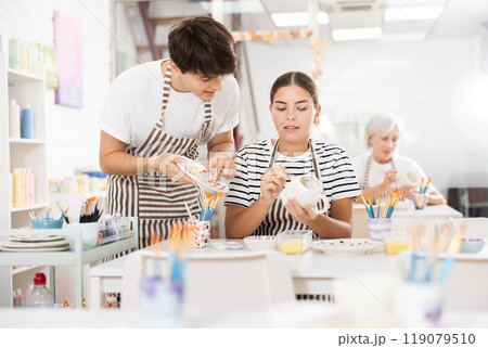 Guy assisting young girl painting ceramic mugs in pottery class 119079510