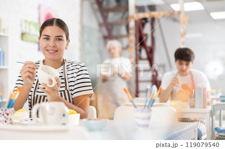 Smiling young female artist painting ceramic cup in pottery workshop Smiling young female artist painting ceramic cup in pottery workshop 119079540