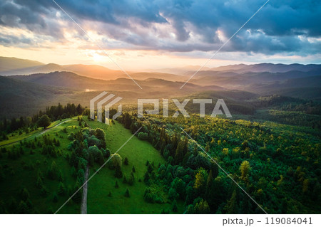 Aerial view breathtaking sunset over forest. Sun's rays break through dramatic clouds, casting golden glow across rolling hills and valleys. Distant mountains creating serene and picturesque scene. 119084041