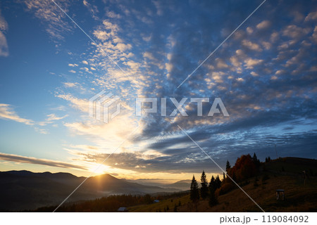 Sun rises over mountains, casting golden rays across landscape. Sky filled with dramatic clouds, creating stunning contrast with clear blue above. Warm light illuminates rolling hills and pine trees. Sun rises over mountains, casting golden rays across landscape. Sky filled with dramatic clouds, creating stunning contrast with clear blue above. Warm light illuminates rolling hills and pine trees. 119084092