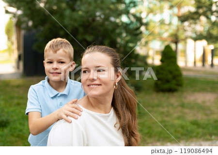 A young boy smiles as he rests his hand on his mother's shoulder, while they stand together outdoors 119086494