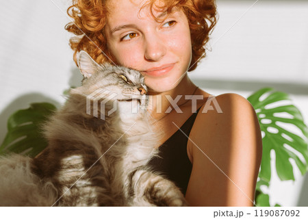 portrait red-haired curly young woman with pet gray fluffy cat in arms, lit by bright sun portrait red-haired curly young woman with pet gray fluffy cat in arms, lit by bright sun 119087092