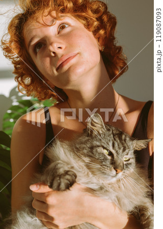 portrait red-haired curly young woman with pet gray fluffy cat in arms, lit by bright sun 119087093