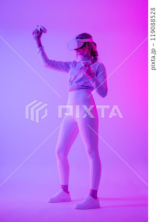 Woman immersed in virtual reality with headset and controllers in neon lights 119088528