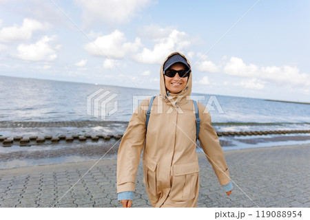 Smiling happy woman walking by Nord sea coast wear beige coat  sunglasses enjoys windy day cloudy sky background. Female person have fun hiking adventure travel at Baltic sea seaside 119088954
