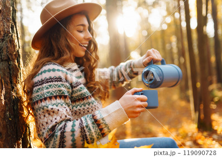 Cute young woman drinks hot drink from thermos and enjoys nature. Autumn landscape. relax concept. Cute young woman drinks hot drink from thermos and enjoys nature. Autumn landscape. relax concept. 119090728