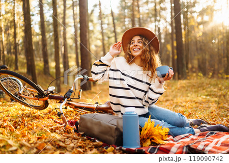Hiker sitting on picnic blanket, drinking hot drink in autumn forest. Woman resting in nature. Hiker sitting on picnic blanket, drinking hot drink in autumn forest. Woman resting in nature. 119090742