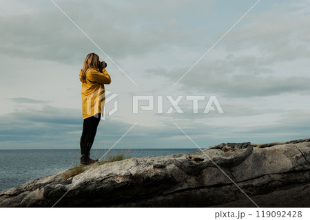 A female in a yellow jacket with a camera stands on a rocky shore, capturing the beauty of nature 119092428