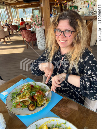 Woman enjoys a meal at a seaside restaurant, with a plate of grilled vegetables and chicken on toast. Bright and open atmosphere, surrounded by palm trees and a scenic view of beach outside Woman enjoys a meal at a seaside restaurant, with a plate of grilled vegetables and chicken on toast. Bright and open atmosphere, surrounded by palm trees and a scenic view of beach outside 119096656