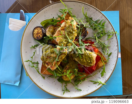 Close-up plate of grilled vegetables and chicken on toast is served in a stylish, bright seaside restaurant. Dish is garnished with fresh arugula and accompanied by a variety of seasonings and Close-up plate of grilled vegetables and chicken on toast is served in a stylish, bright seaside restaurant. Dish is garnished with fresh arugula and accompanied by a variety of seasonings and 119096663