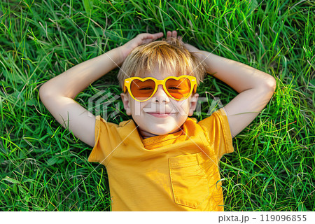 Happy stylish child lying on spring green grass. 4-5 years old boy lying back on perfect lawn top view. Kid wearing yellow t-shirt, red heart shaped sunglasses outdoors in a backyard. Summer activity 119096855