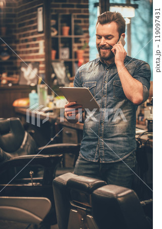 our appointment is on Monday! Cheerful young bearded man talking on mobile phone and looking at digital tablet while standing at barbershop 119097431