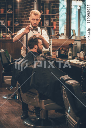 Barber at work. Full length of young bearded man getting haircut by hairdresser while sitting in chair at barbershop 119098925