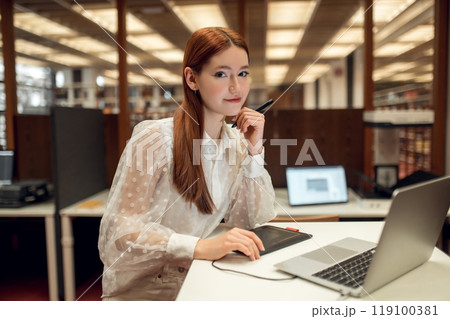 Long-haired pretty ginger girl working on a laptop in the library 119100381