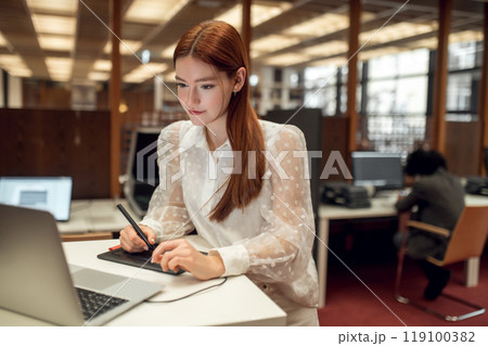 Long-haired pretty ginger girl working on a laptop in the library Long-haired pretty ginger girl working on a laptop in the library 119100382