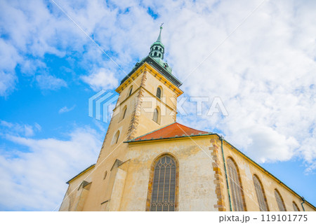 The Church of the Assumption of the Virgin Mary showcases stunning late Gothic architecture against a bright blue sky in Most, Czechia. Its tower reaches towards the clouds, captivating visitors. 119101775