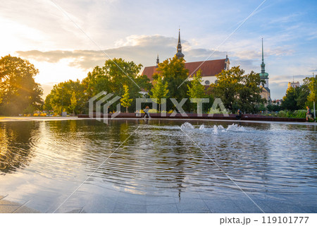 As evening settles, sunlight illuminates the Moravian Square fountain in Brno. The shimmering water reflects nearby trees and historic buildings, creating a tranquil atmosphere. 119101777