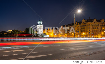 A long exposure photograph captures the vibrant lights of Prague at dusk, showcasing the Sitkov Water Tower and the Masaryk Embankment. Traffic streaks illuminate the road in front of the buildings. 119101794