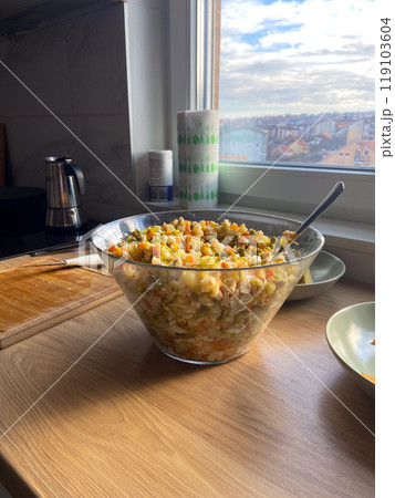 Clear glass bowl filled with a colorful vegetable salad sits on a sunlit kitchen counter. Mix of ingredients reflects the freshness and simplicity of home-cooked meals enjoyed in a cozy kitchen 119103604